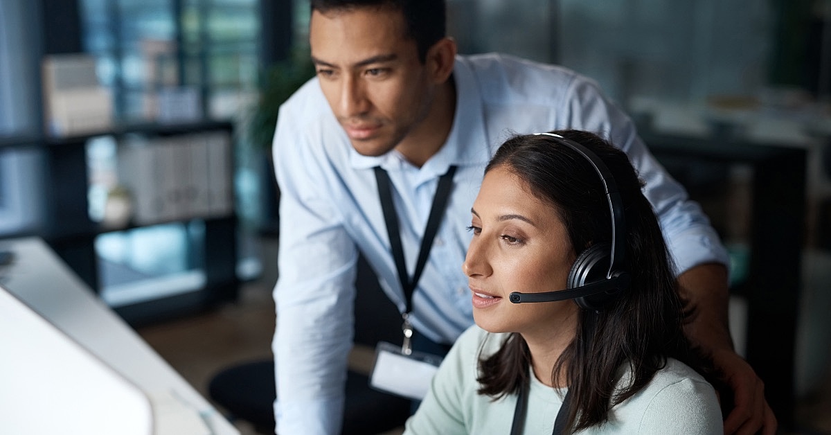 a imagem mostra uma profissional de contact center durante um atendimento e um colega de trabalho ao seu lado observando.