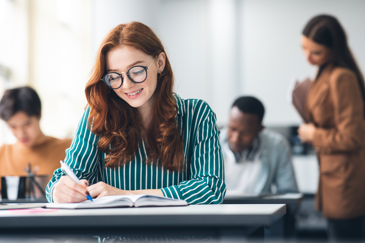 Education, High School, Learning and People Concept. Portrait of beautiful ginger female student in glasses writing test in classroom, sitting at table with group of classmates in blurred background