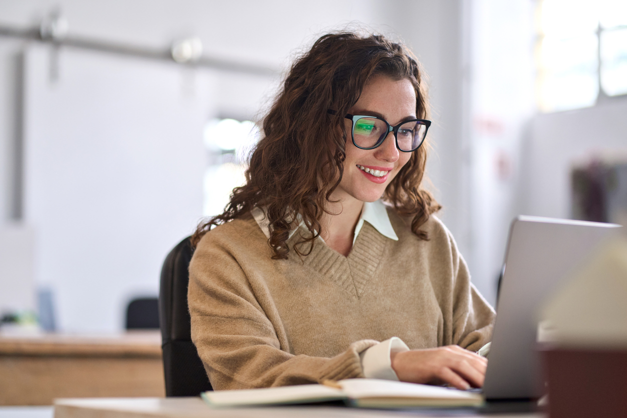 Mulher jovem feliz profissional de negócios sentado na mesa trabalhando no laptop no escritório corporativo. Aluna sorridente usando tecnologia de computador aprendendo on-line, fazendo pesquisas na web.